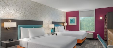 A hotel room with two beds, a nightstand with a lamp, a wall-mounted TV, and a window with a view of greenery.
