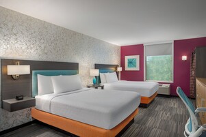 Habitación de hotel con dos camas, una mesita de noche con lámpara, un televisor montado en la pared y una ventana con vistas a zonas verdes.