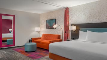 Quarto de hotel com uma cama grande, um sofá laranja, um banquinho azul, um espelho e um quadro na parede.
