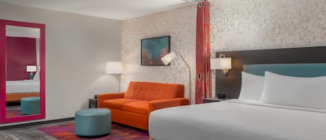 Una habitación de hotel con una cama grande, un sofá naranja, un reposapiés azul, un espejo y un cuadro en la pared.