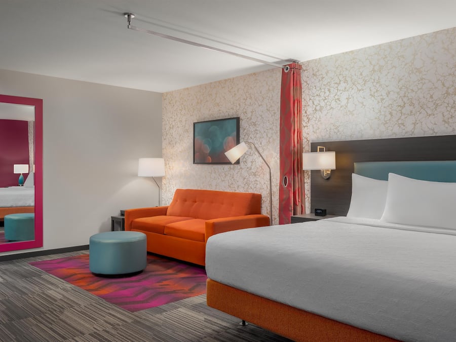 Una habitación de hotel con una cama grande, un sofá naranja, un reposapiés azul, un espejo y un cuadro en la pared.