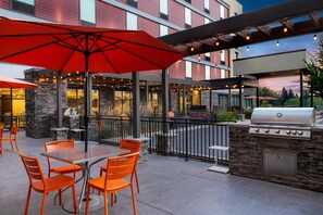 An outdoor patio with a table and chairs, a grill, and a view of a building. - Home2 Suites by Hilton Portland Hillsboro (Hillsboro)