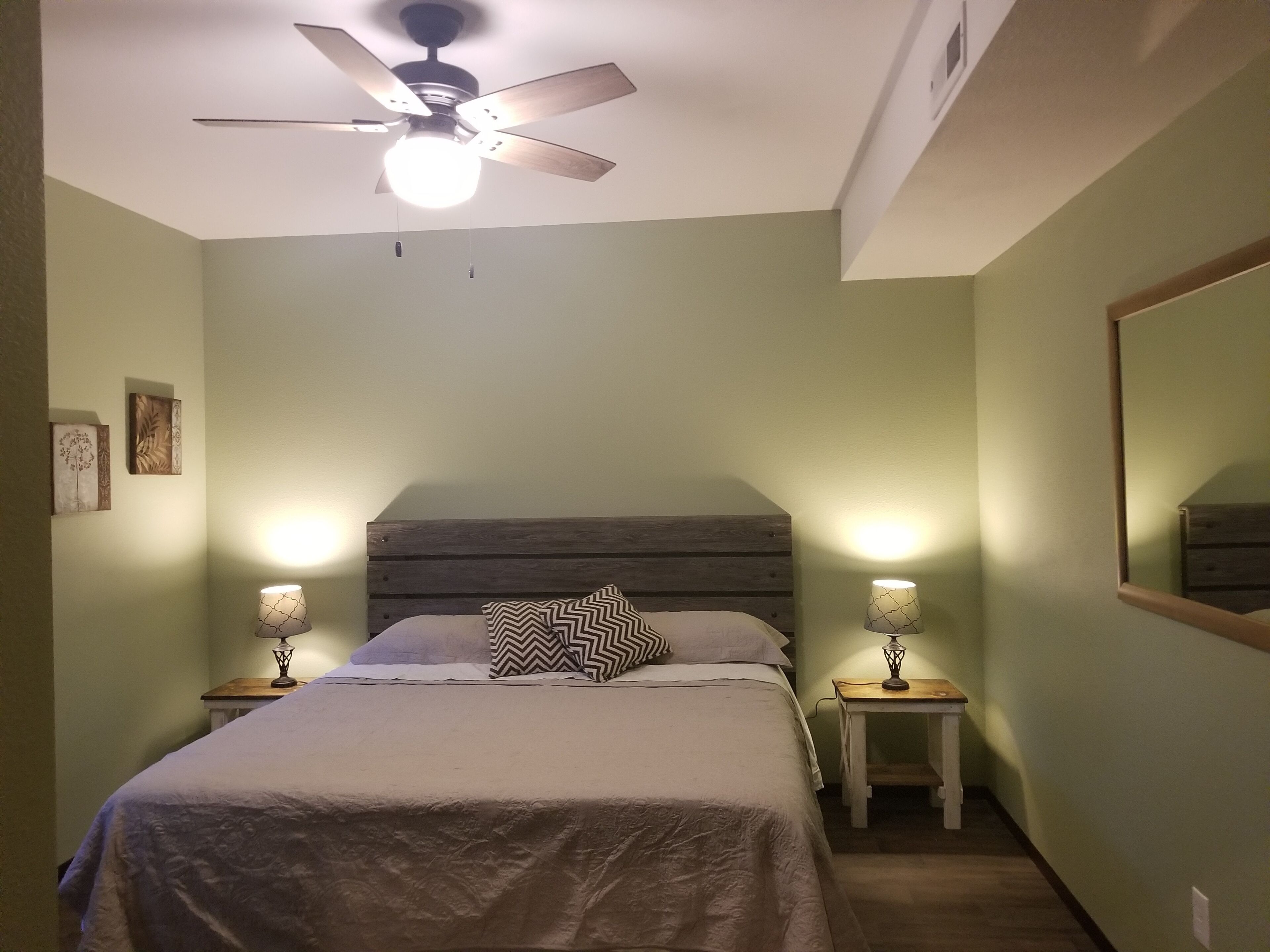 Une chambre à coucher avec un lit, deux tables de chevet avec des lampes, un ventilateur de plafond et un miroir au mur.