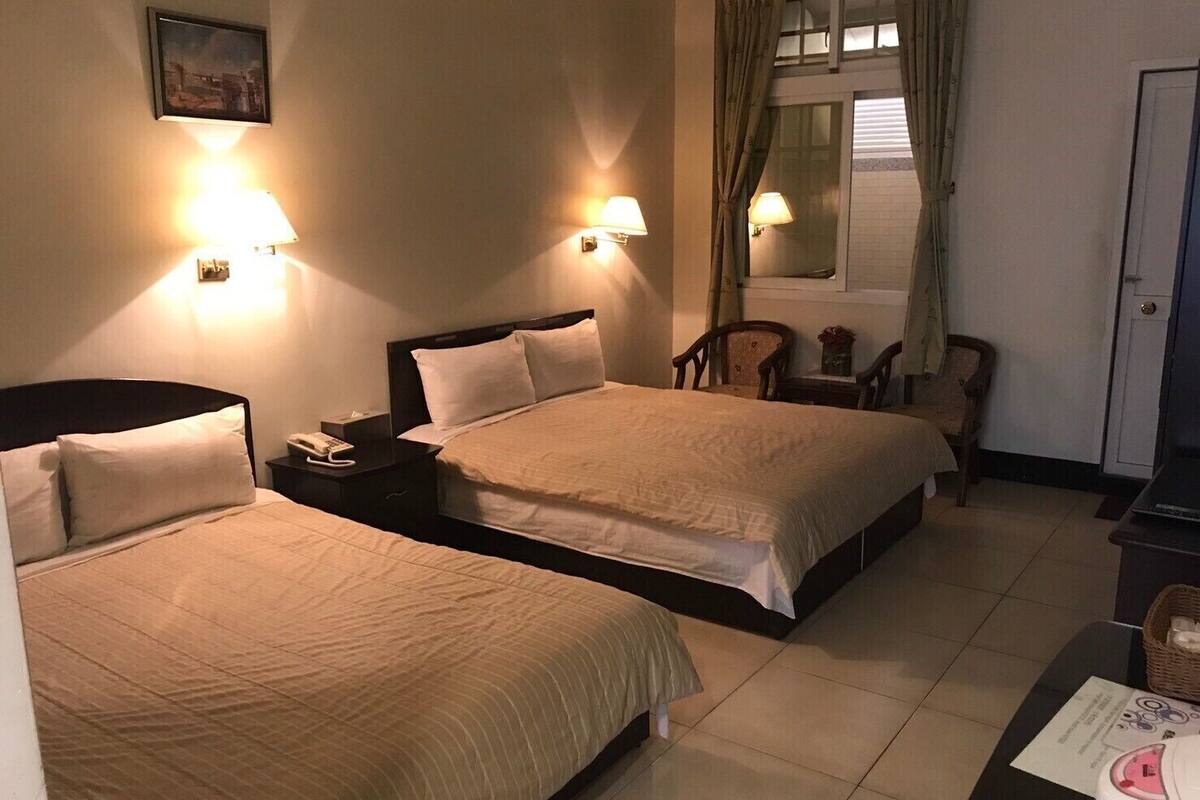 A hotel room with two beds, bedside lamps, a small table with a telephone, a chair, and a window with curtains.