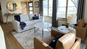 A living room with a sofa, two armchairs, a coffee table, a side table with a lamp, a mirror, and a view of the beach. - Summer Sand & Fun Beachfront Vacation Traverse City In Cottage (Traverse City)