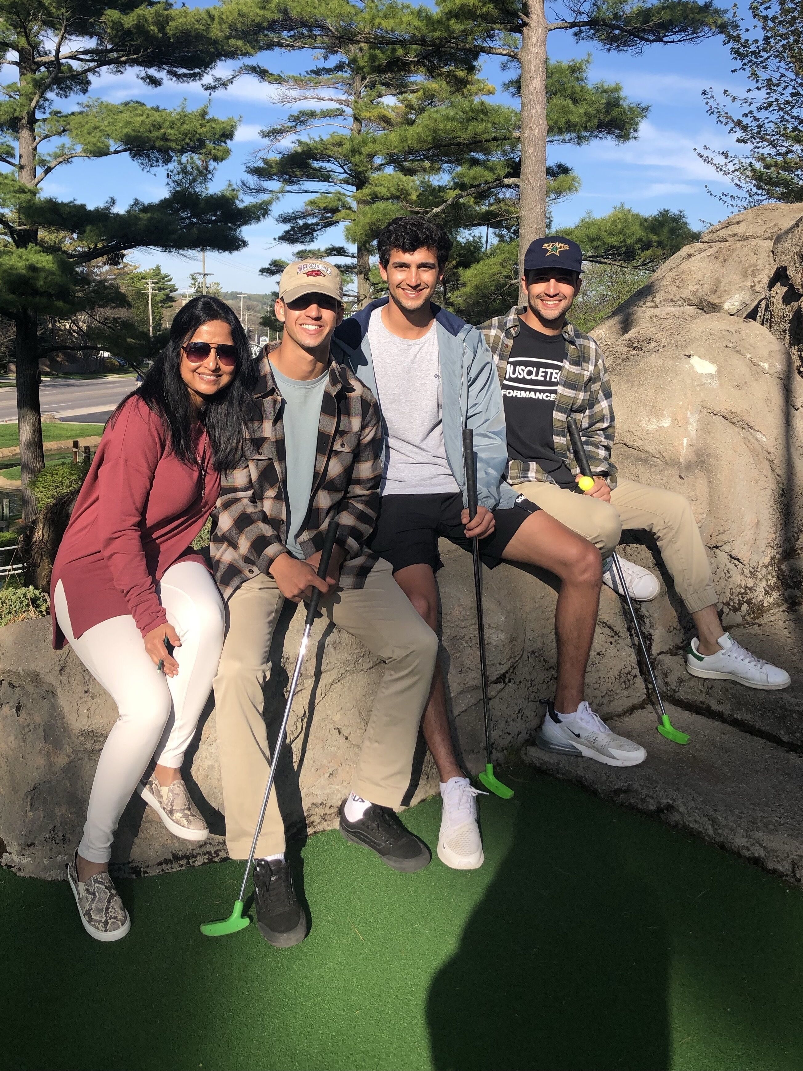 Un grupo de personas sentadas sobre una formación rocosa, con un palo de golf en la mano, rodeadas de árboles y un cielo despejado.