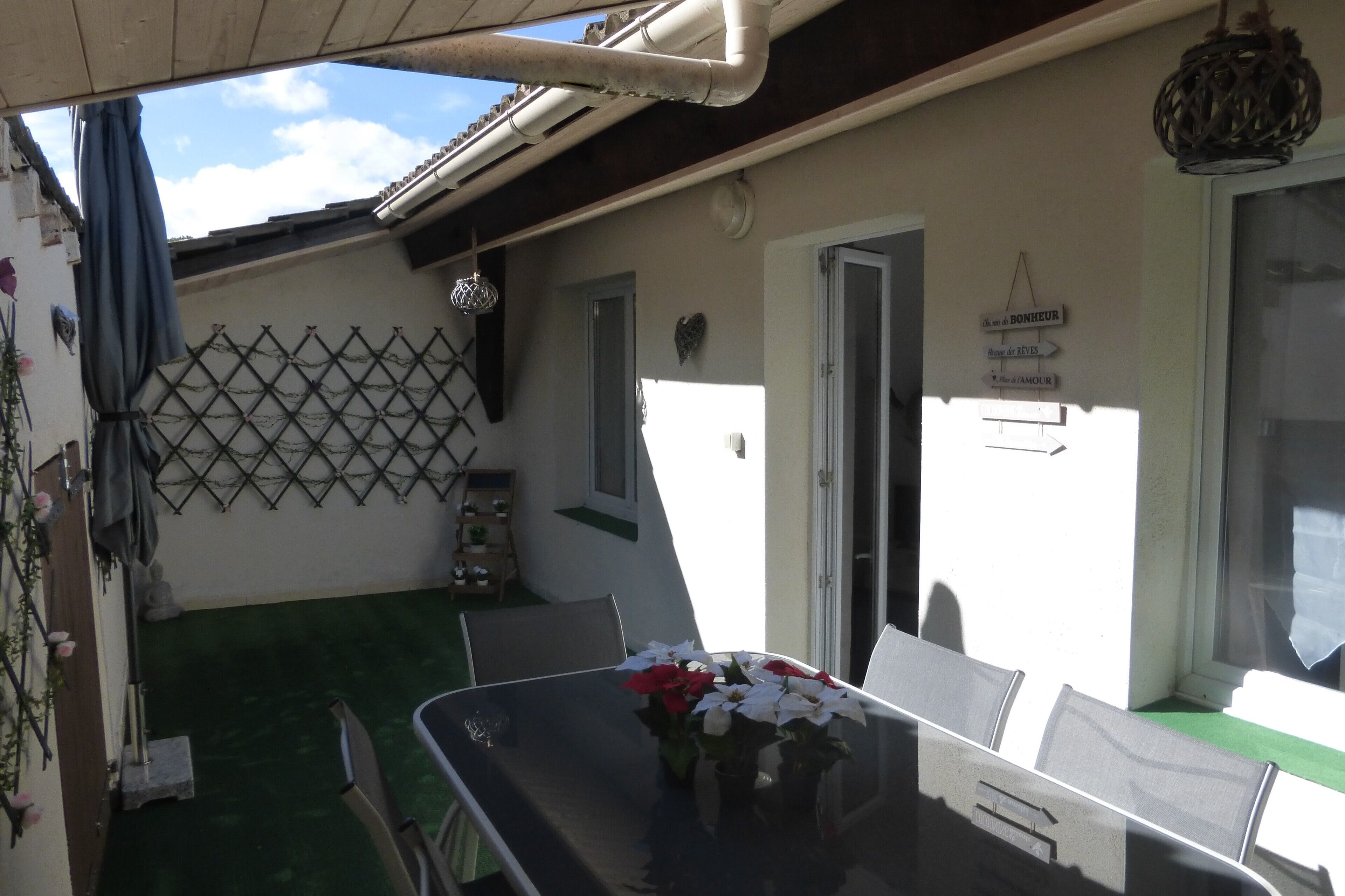 Un patio con una mesa de vidrio, sillas y un jarrón de flores. Se observa una pared enrejada y una puerta a la derecha.