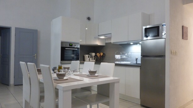A modern kitchen with white cabinetry, a stainless steel refrigerator, an oven, and a microwave. There is a dining table with chairs and a bowl on it.