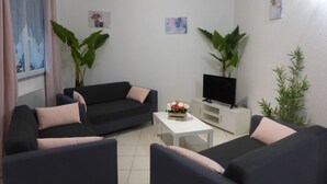 A living room with a dark sofa set, a white coffee table, and a flat-screen TV on a white cabinet. There are two large potted plants, one on each side of the room.