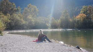 Une personne assise sur une plage de galets, au bord d’une rivière paisible, entourée d’arbres et sous un ciel dégagé.