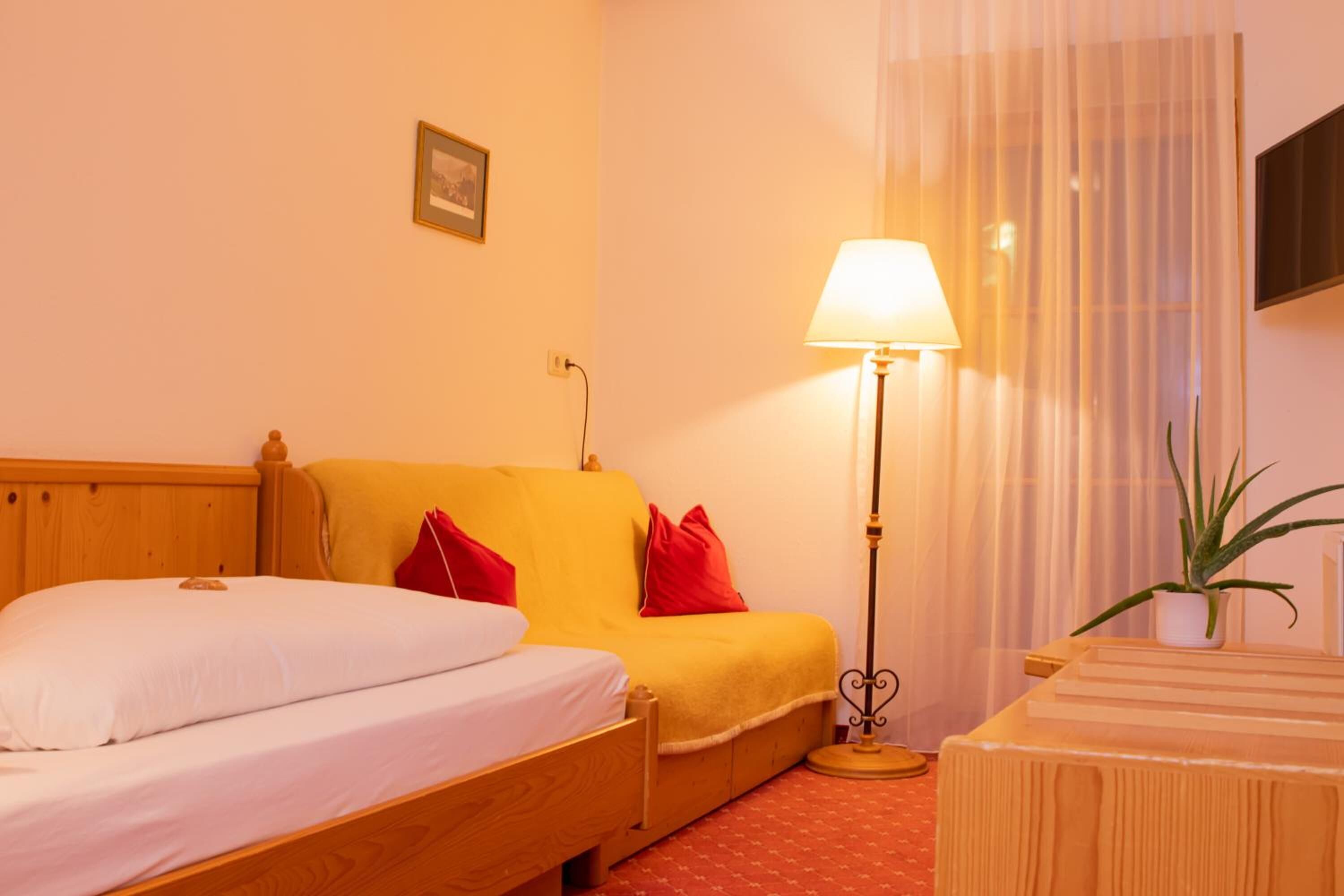 A hotel room with a wooden bed, a yellow sofa, a bedside table, a lamp, and a potted plant.