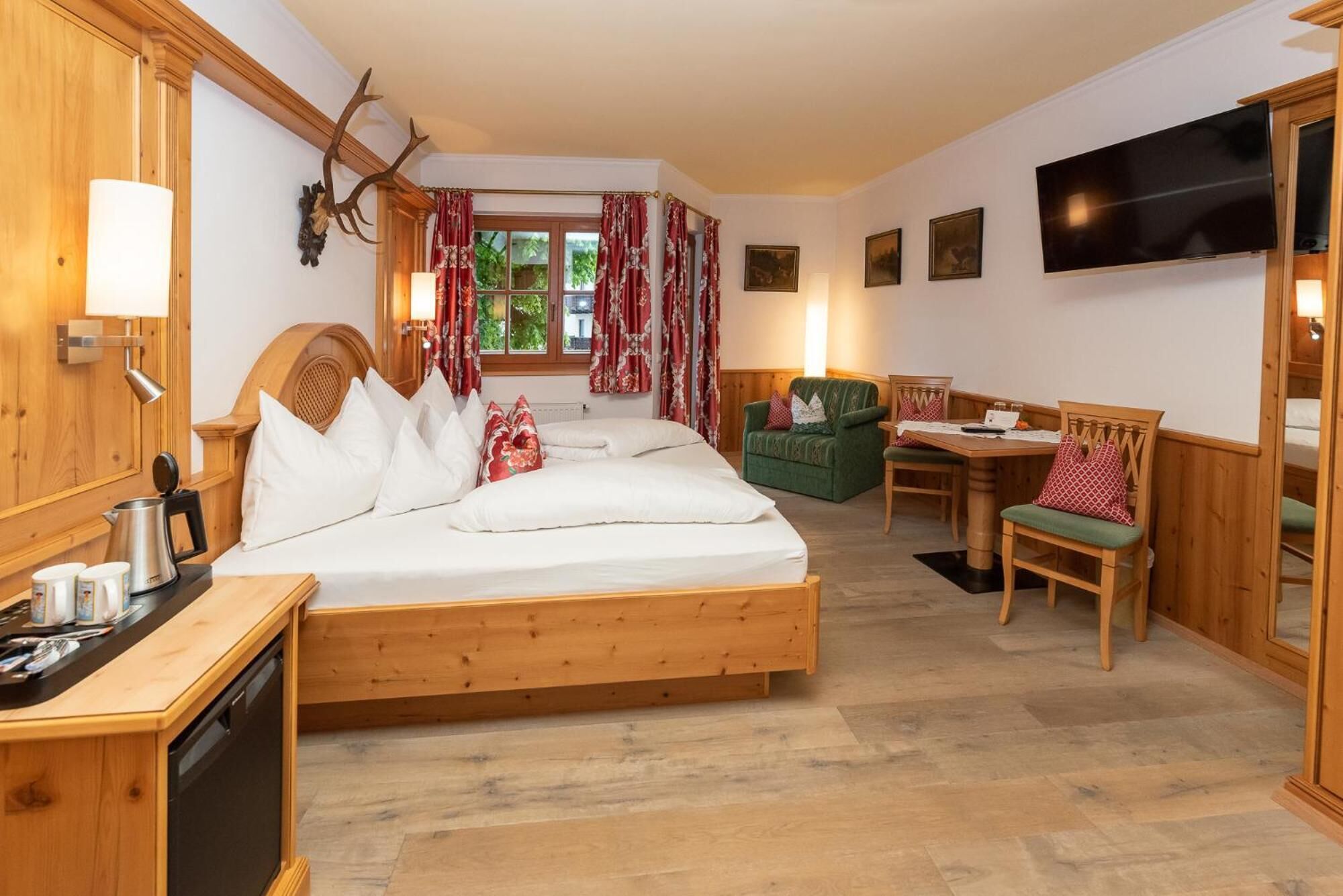 A hotel room with a wooden bed, a desk with a kettle, a green armchair, and a TV on the wall.