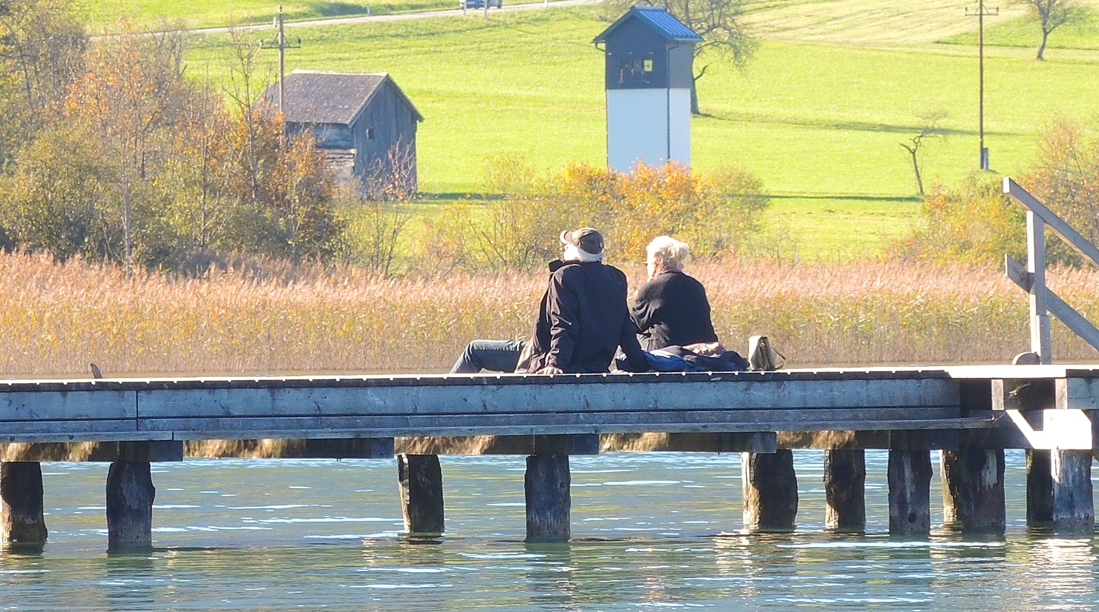 A wooden pier extending into a body of water with two people sitting on it. In the background, there is a grassy field with a small building and a utility pole.