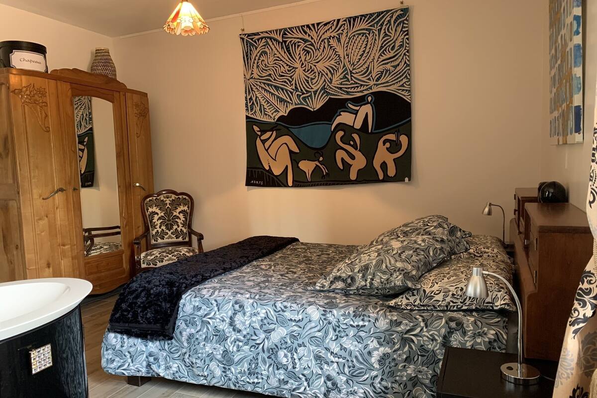 A bedroom with a bed, a chair, a wardrobe, a bedside table, and a wall hanging.
