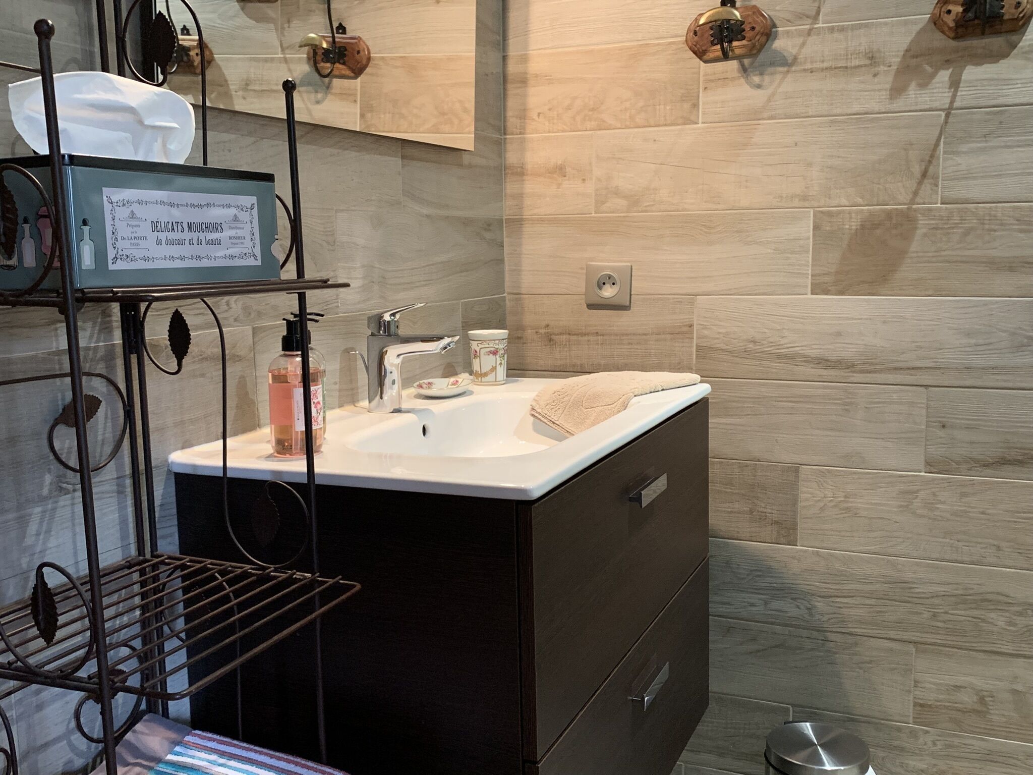 A modern bathroom with a dark cabinet, a white sink, and a mirror above it. There is a shelf with toiletries and a towel on the sink.