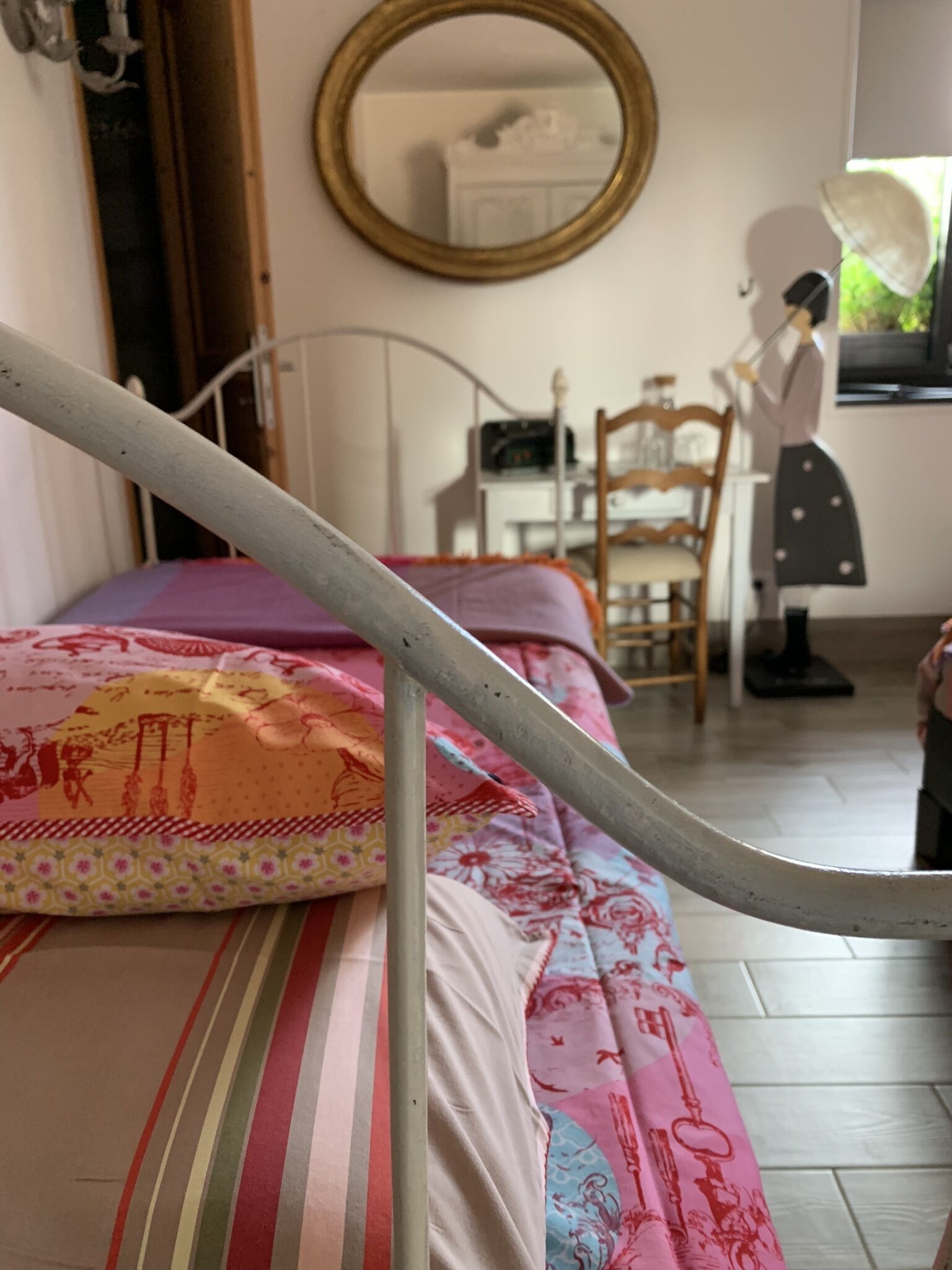 Un lit avec un cadre en métal, une couvre-lit colorée, une chaise en bois, un bureau avec un ordinateur et un miroir accroché au mur.