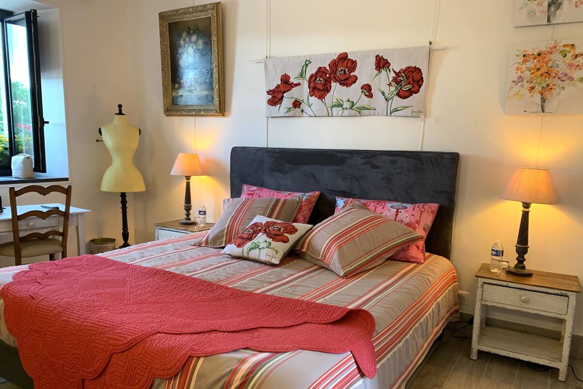 A neatly made bed with striped bedding and a pink throw, flanked by two bedside tables with lamps, a dresser, and a painting of flowers on the wall.