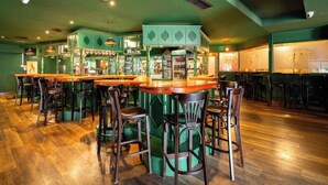 A bar area with wooden tables and chairs, a counter with a green backdrop, and a variety of bottles and glasses. - Santa Maria Aparthotel (Adeje)
