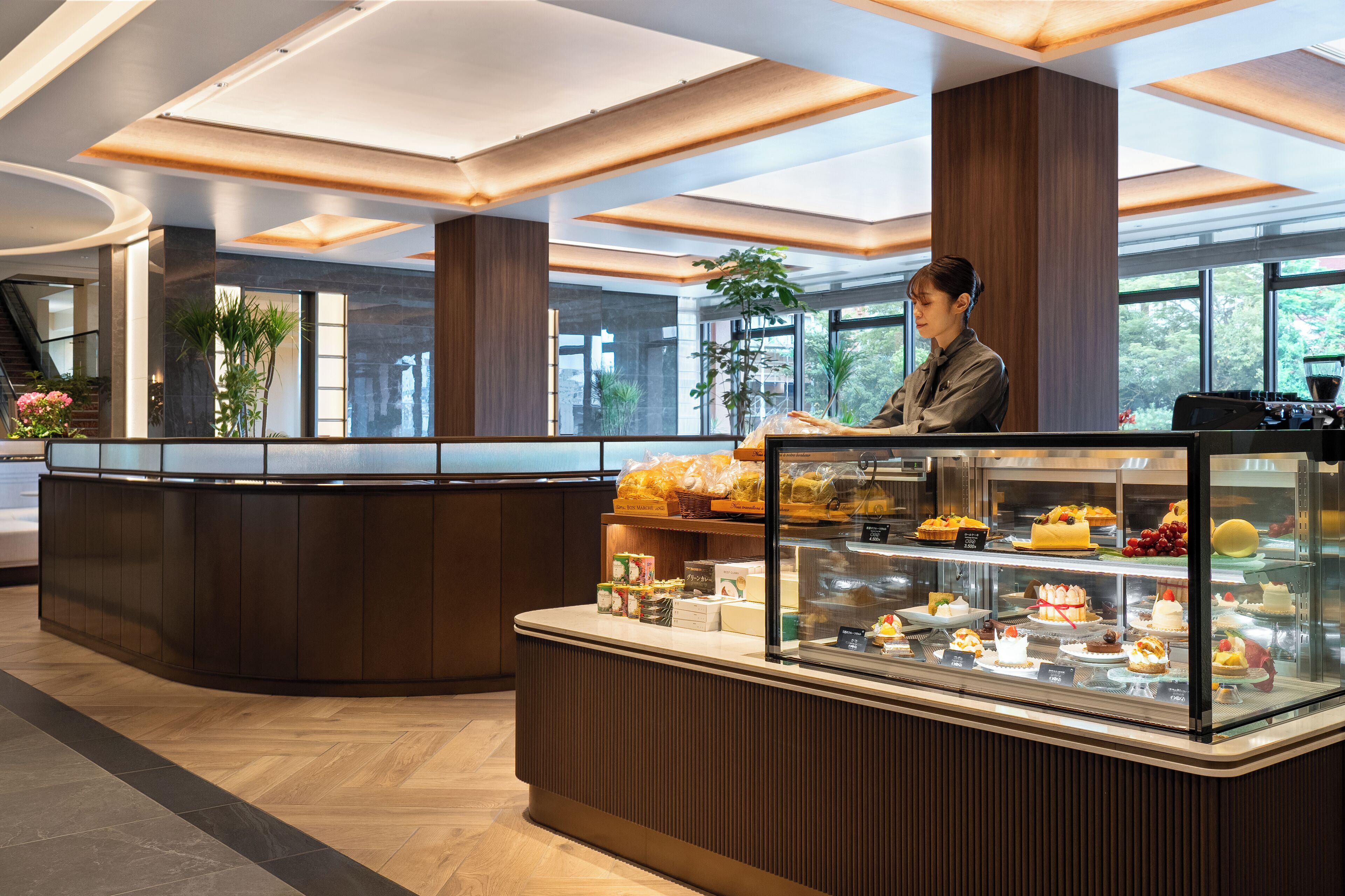 A modern reception area with a curved counter, a glass display case with pastries, and large windows offering a view of greenery outside.