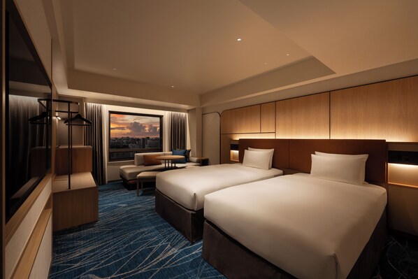 Habitación de hotel con dos camas, una zona de estar, un ventanal grande con vista a la ciudad y un cabecero de madera.