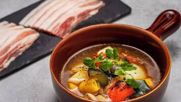 A bowl of soup with vegetables and herbs, a wooden spoon, and a black plate with sliced meat in the background.