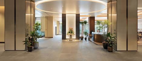 A spacious, modern lobby with a curved ceiling, large potted plants, and a reception desk.