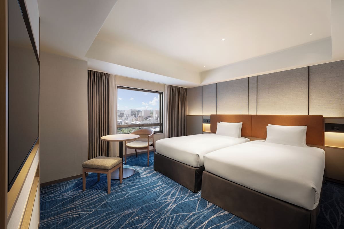 Ett hotellrum med två sängar, ett litet bord, en stol och ett stort fönster med utsikt över staden.