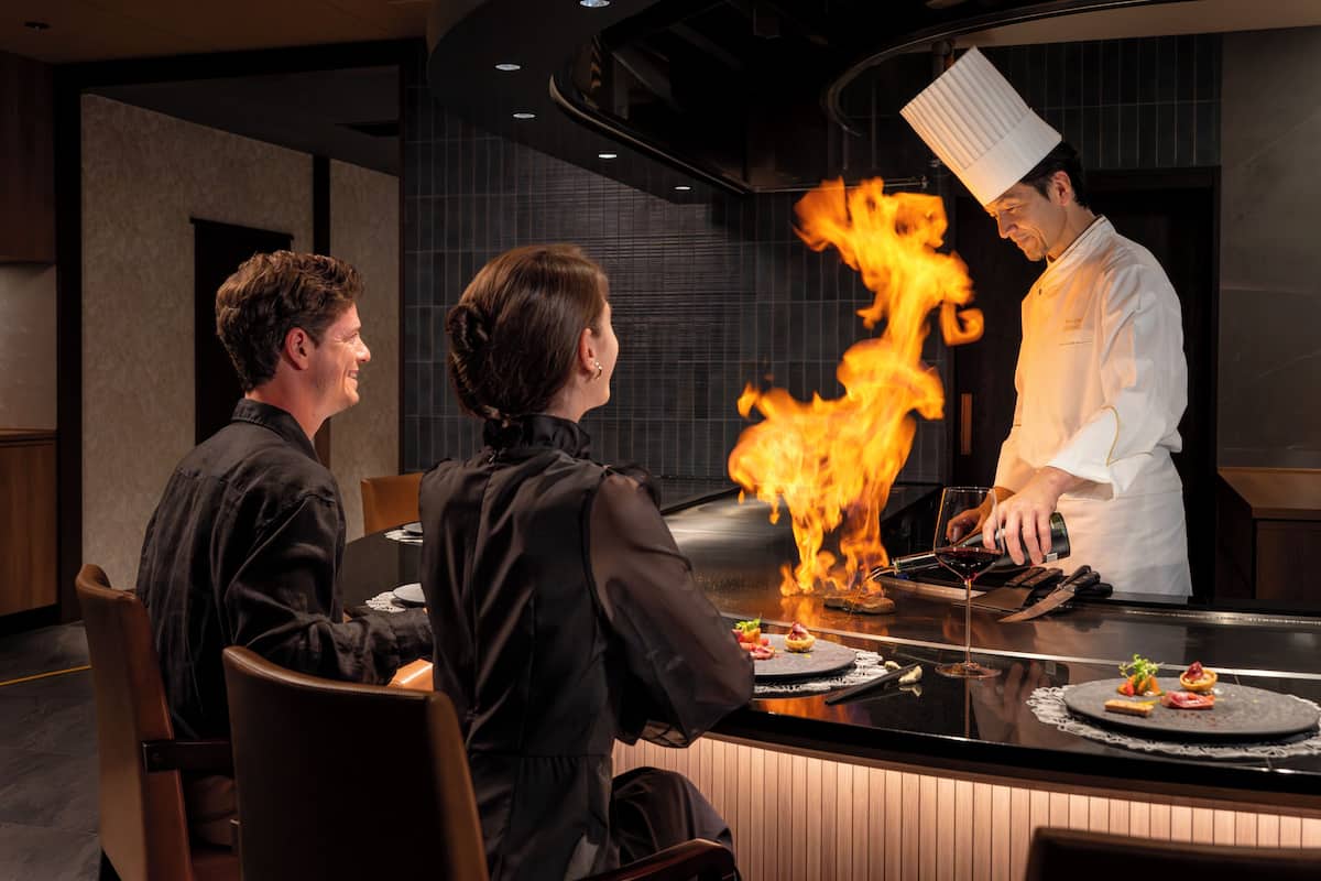 En kock som tillagar en rätt med hjälp av flambétekniken vid en bardisk i restaurangen, medan gästerna sitter vid borden.