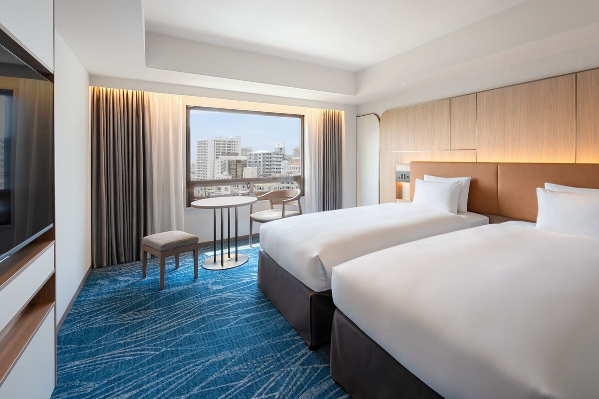 Habitación de hotel con dos camas, vista a la ciudad, una mesa de comedor y un televisor.