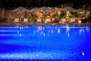 Piscina con sillas de descanso y sombrillas, rodeada de rocas y plantas.