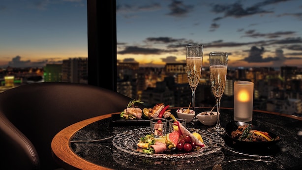 A table set with a glass of champagne, a candle, and a variety of appetizers against a cityscape view. - Okinawa Harborview Hotel (Naha)