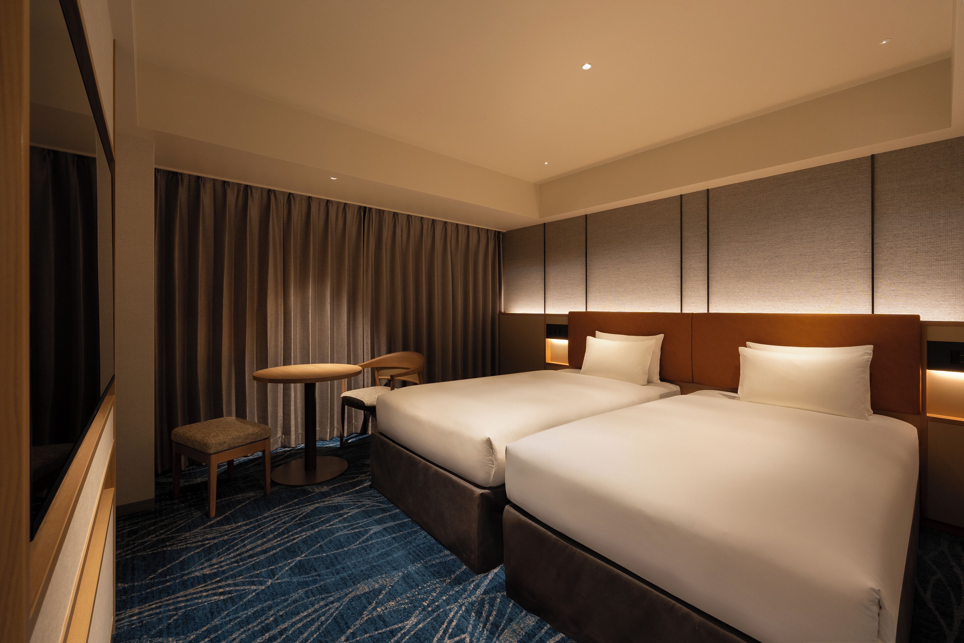 Habitación de hotel con dos camas, un escritorio, una silla y un ventanal con cortinas.