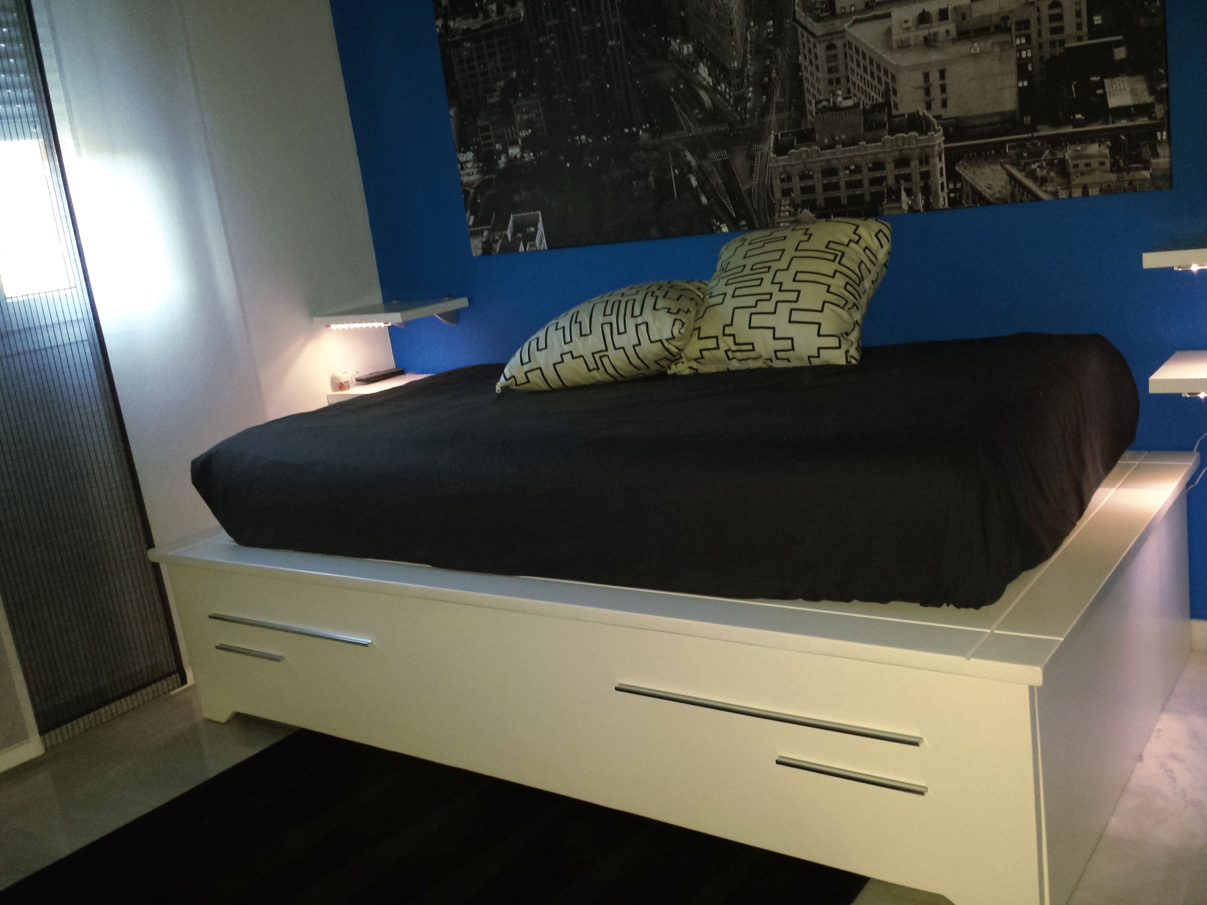 Een moderne slaapkamer met een bed, zwart-wit gestreepte kussens, een behang met een stadsgezicht en een witte nachtkastje met een lamp.