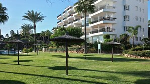 Een verzorgd gazon met rieten parasols, palmbomen en een modern gebouw met balkons op de achtergrond.