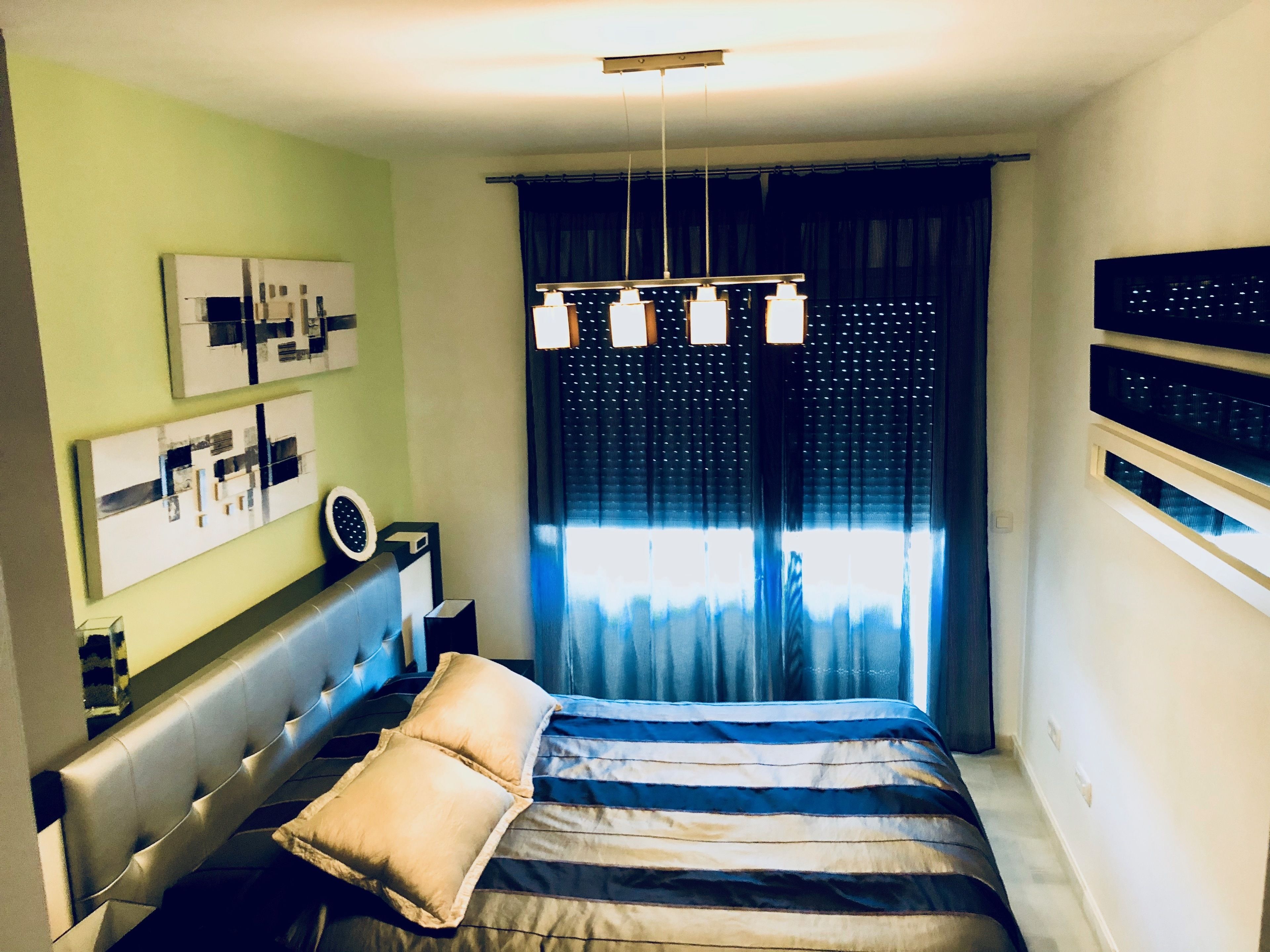 Un dormitorio con cama, cabecera, mesita de noche, lámpara y ventana con cortinas.