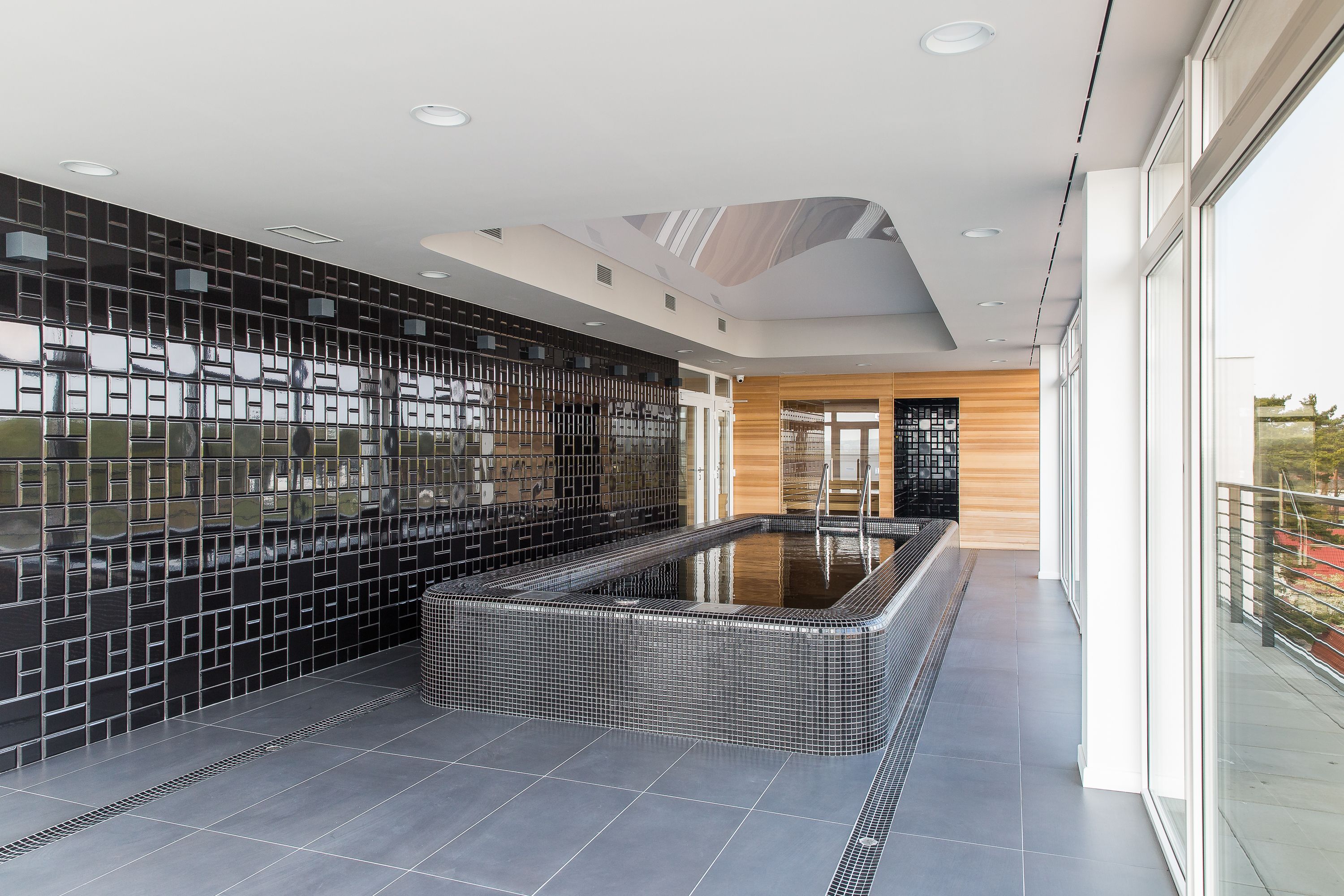 A modern indoor swimming pool with a tiled floor and walls, surrounded by a balcony with glass railings.