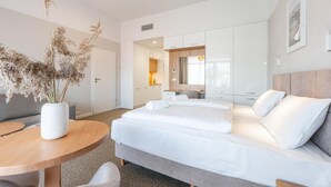 Une chambre d’hôtel moderne avec un lit, une table ronde en bois, un vase contenant des plantes séchées et un grand miroir.