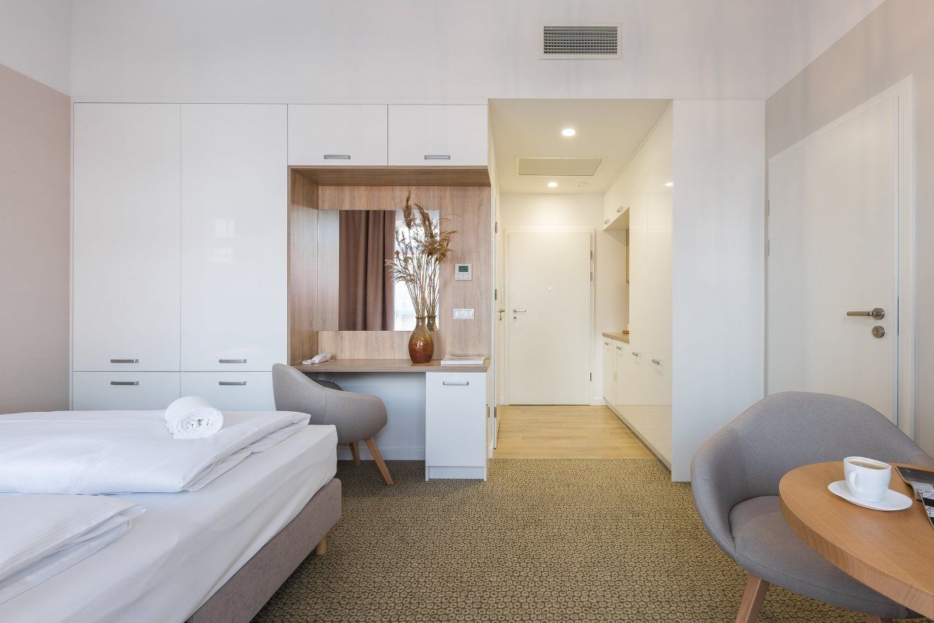A hotel room with a bed, a desk, a chair, a small table with a cup, and a door leading to another room.
