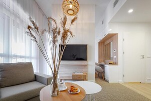 A modern living room with a sofa, a coffee table with pastries, a vase with dried plants, and a TV mounted on the wall.