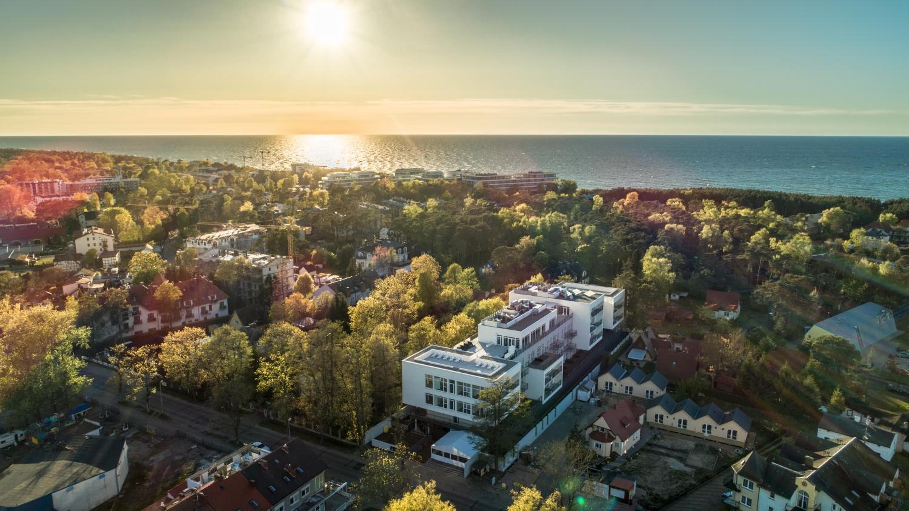 Vista aérea de un pueblo costero con una mezcla de edificios residenciales y comerciales, un edificio moderno destacado en el centro, y una masa de agua al fondo.