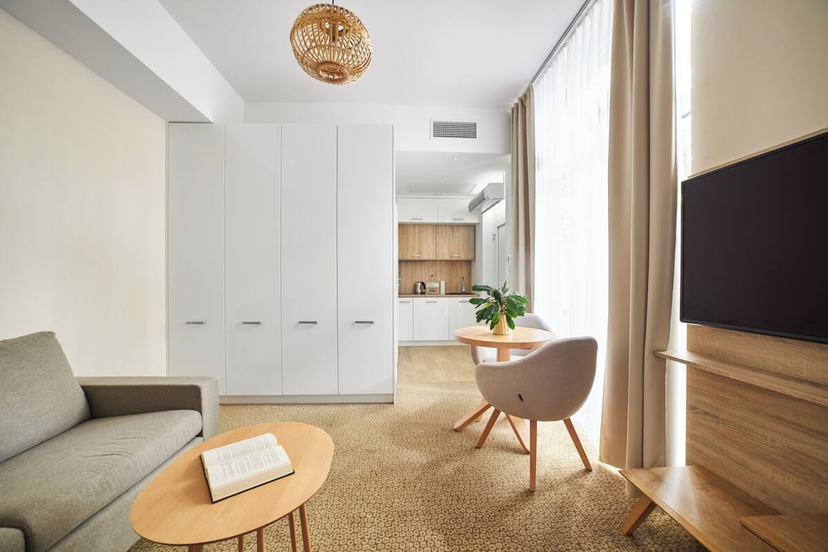 Ein modernes Hotelzimmer mit einer Couch, einem runden Tisch mit einem Buch, einem Fernseher, einem Essbereich mit einer Pflanze und einer Küche im Hintergrund.