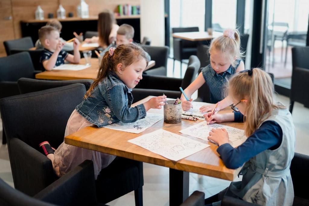 A group of children engaged in drawing activities at a table in a modern indoor setting with black chairs and wooden tables.