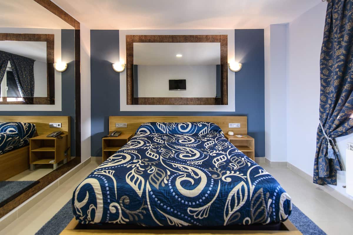 Habitación de hotel con una cama grande, un espejo sobre la cama, mesitas de noche y una ventana con cortinas.