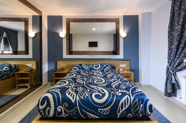 Habitación de hotel con una cama grande, un espejo sobre la cama, mesitas de noche y una ventana con cortinas.