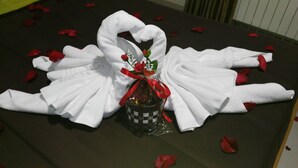 A bed with a towel arranged in a heart shape, a small gift basket, and red rose petals scattered on the bed. - Motel Cancún Avilés (Gozon)