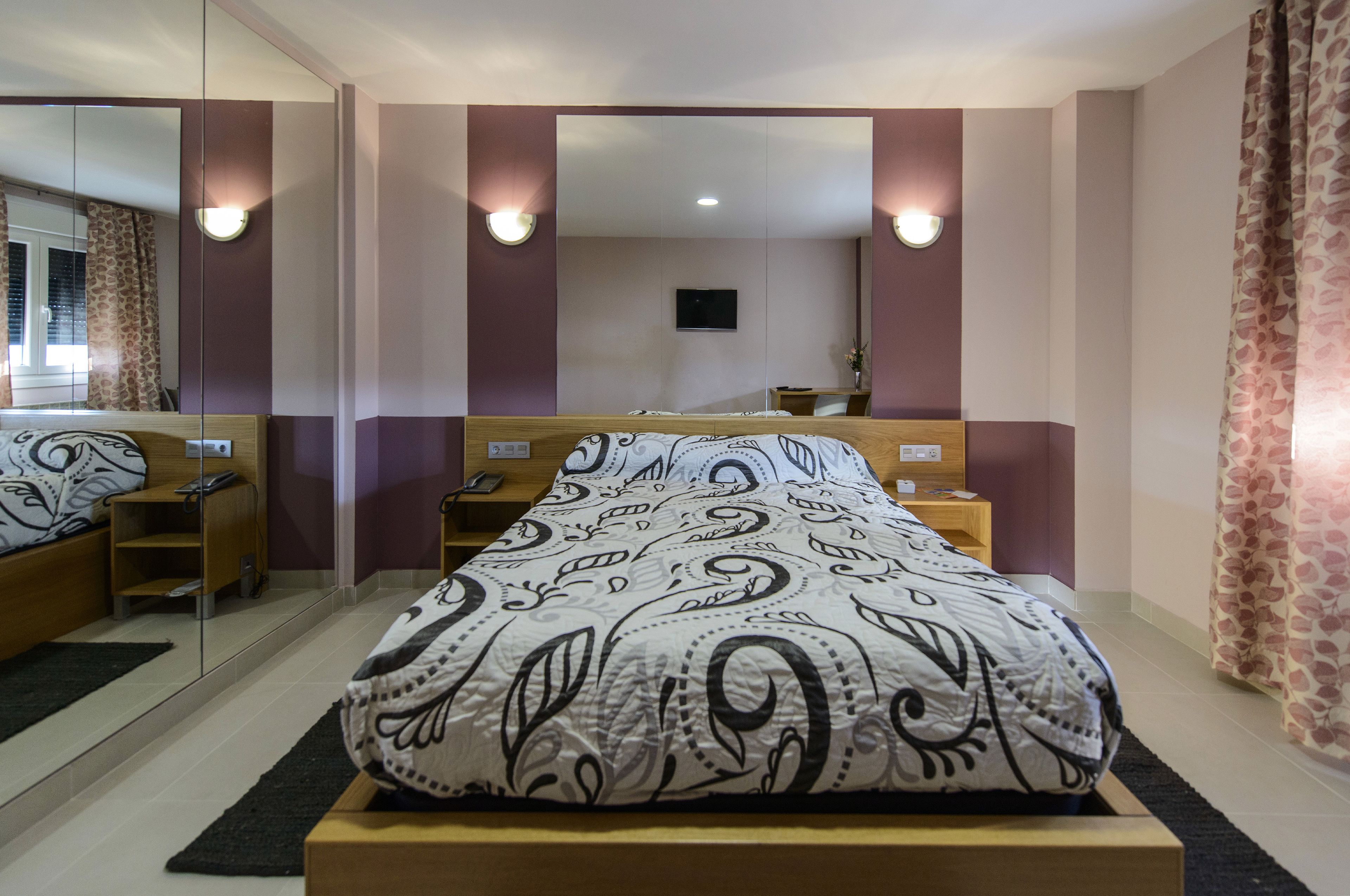 Een slaapkamer met een bed, nachtkastje, spiegel en een tv aan de muur.