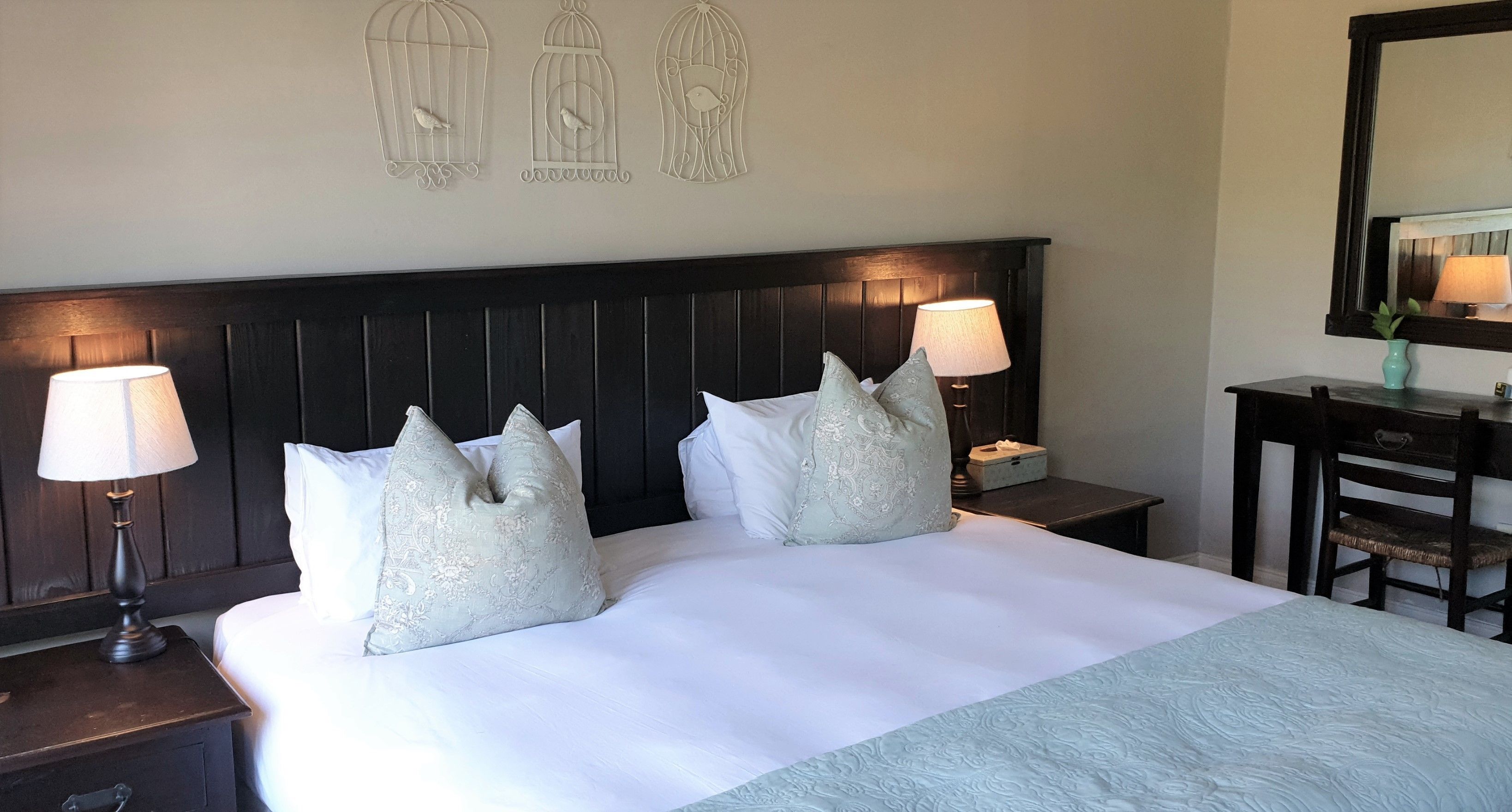 Een netjes opgemaakt bed met twee decoratieve kussens, een nachtkastje met een lamp en een spiegel aan de muur.