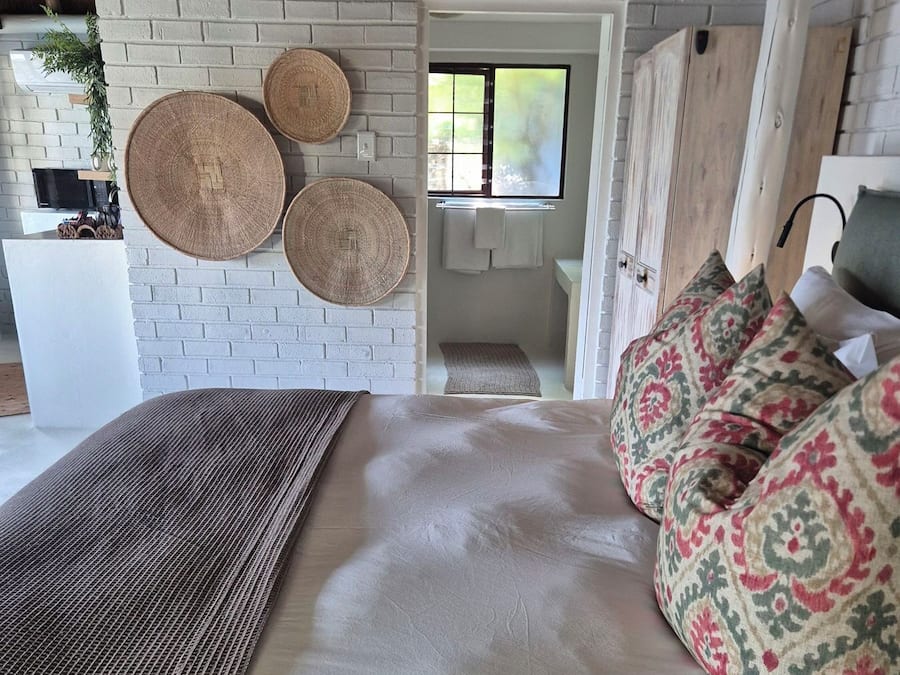 A bedroom with a bed, a woven rug, a patterned pillow, and a brick wall.