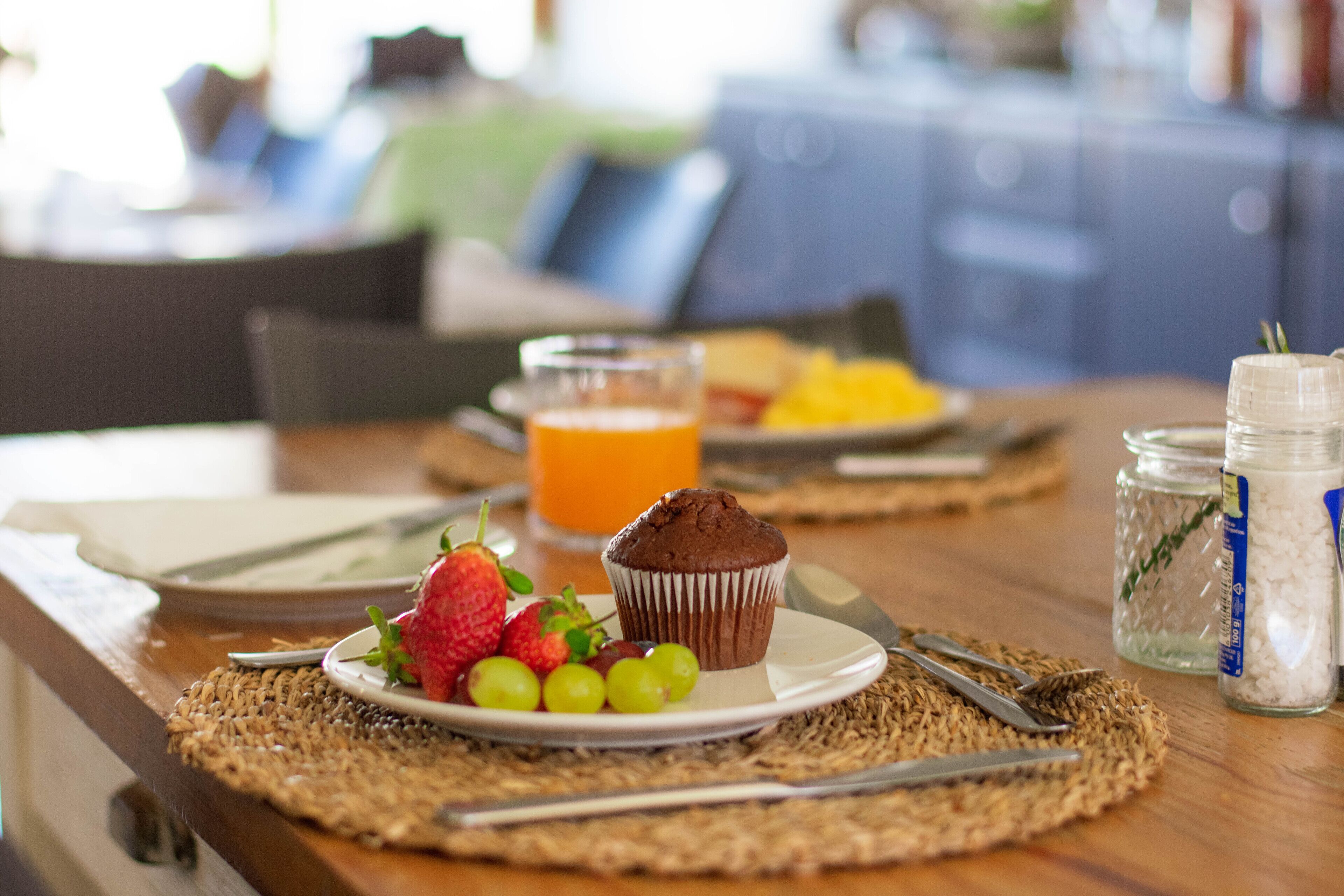 A breakfast table with a plate of strawberries, grapes, and a muffin, a glass of orange juice, and a jar of sugar.