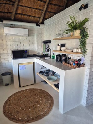 A compact kitchen with a white brick wall, a thatched ceiling, a built-in microwave, a small refrigerator, and a wooden countertop with various kitchen items.